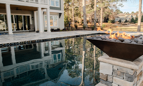 A modern backyard with a swimming pool, a stone fire pit with flames burning, a patio area, and a large two-story house surrounded by trees and landscaped grass.