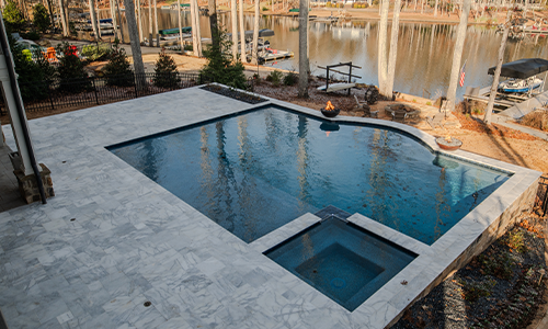 A modern backyard pool with a connected hot tub, surrounded by a stone patio. The pool overlooks a scenic lake with docks, boats, and tall trees in the background.