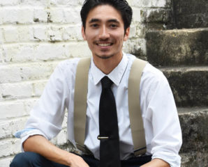 John Ryan Lechner smiling and sitting on a brick staircase. He needs a haircut and to tuck in his shirt in the back more