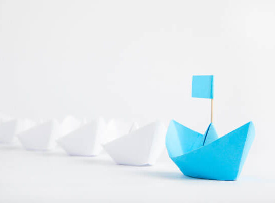 blue paper boat in front of a line of white paper boats