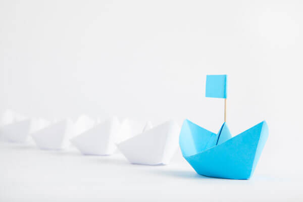blue paper boat in front of a line of white paper boats