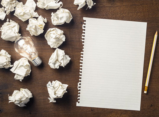 crumpled pieces of paper are sitting on a table next to a clean sheet of paper and a lightbulb