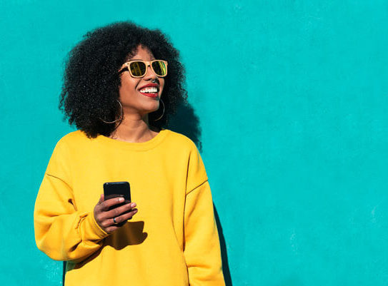 a smiling young woman is wearing sunglasses and using her mobile smart phone