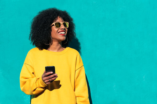 a smiling young woman is wearing sunglasses and using her mobile smart phone