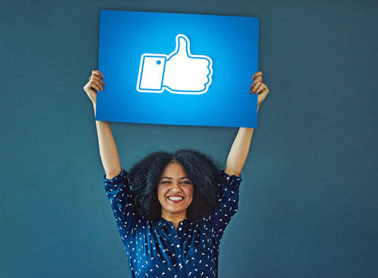 smiling young woman triumphantly holds a sign above her head that has a 'thumbs up' icon on it