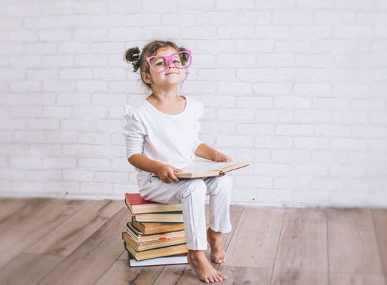 young girl with funny looking, large glasses sits on a pile of books while holding a book in her hand