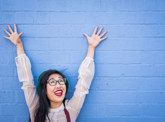 smiling young woman cheerfully raises her arms in the air in front of a painted blue wall