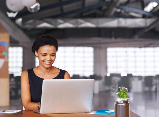 a young, smiling woman using a laptop and notebook to complete a task