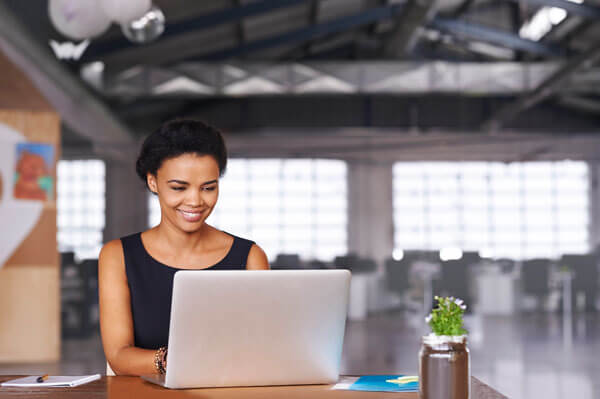 a young, smiling woman using a laptop and notebook to complete a task