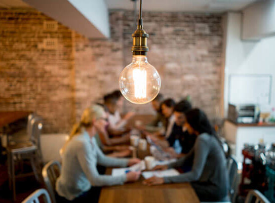 lightbulb in focus hanging over a table with young people working on a project together