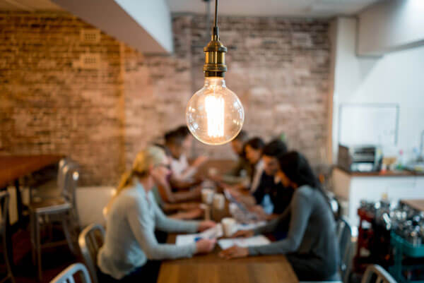 lightbulb in focus hanging over a table with young people working on a project together