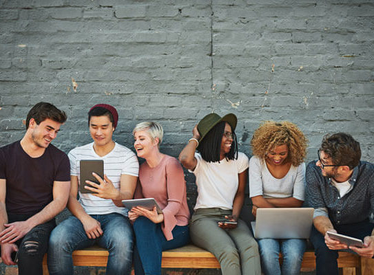 group of young adults sit on a bench together and are holding smart phones, tablets, and laptops