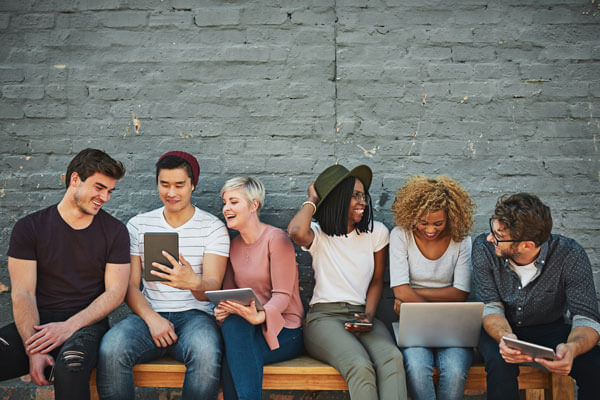 group of young adults sit on a bench together and are holding smart phones, tablets, and laptops