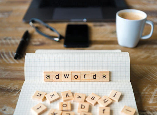 scrabble board game letters spell out 'adwords' and sit on a table in front of a phone, laptop, and cup of coffee