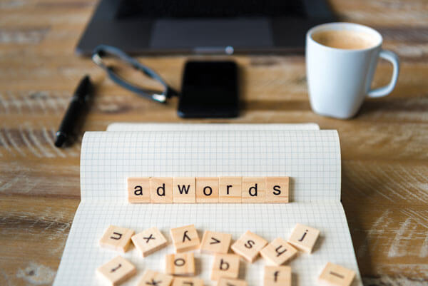 scrabble board game letters spell out 'adwords' and sit on a table in front of a phone, laptop, and cup of coffee