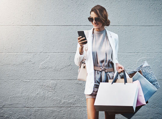 fashionable young woman carrying several shopping bags smiles and looks at her phone