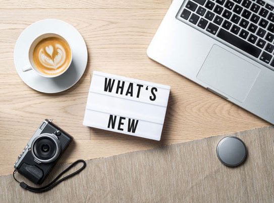 a sign that says 'whats new' placed on a table next to a laptop, a cup of coffee, and a camera