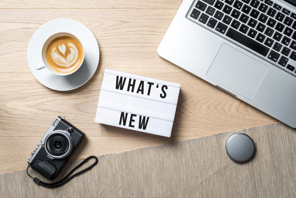a sign that says 'whats new' placed on a table next to a laptop, a cup of coffee, and a camera