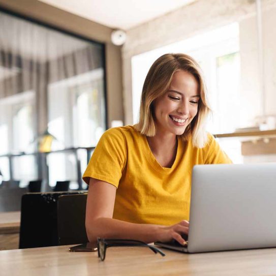 Photo of joyful nice woman using laptop and smiling while sitting at table in open-plan office