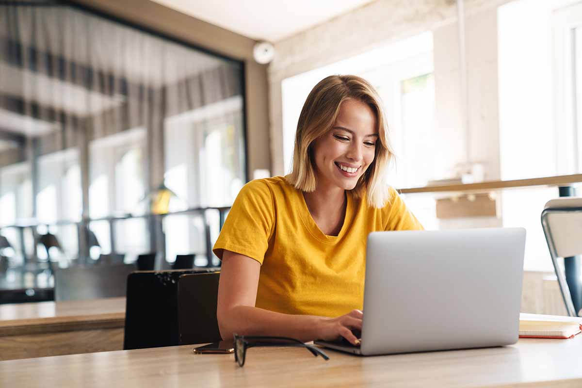 Photo of joyful nice woman using laptop and smiling while sitting at table in open-plan office