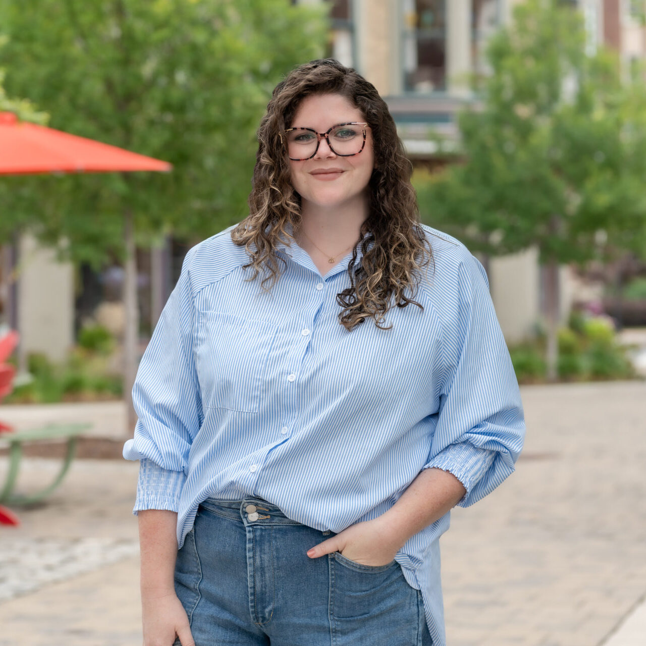 A woman with curly brown hair and glasses stands outdoors, smiling. She wears a light blue striped shirt and jeans with one hand in her pocket. The background features trees, an orange umbrella, and blurred buildings.