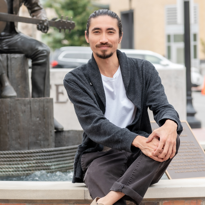 A man with long dark hair tied back, wearing a white shirt and dark cardigan, sits cross-legged on a low wall in front of a statue and fountain, with buildings and a parked car in the background.
