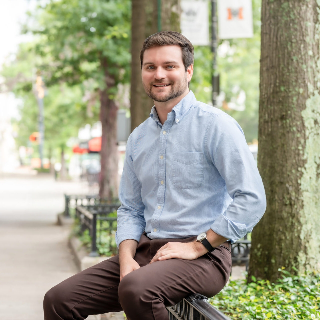 A man with short brown hair and a beard, wearing a light blue button-down shirt and brown pants, sits on a park railing, smiling. Trees and greenery line the sidewalk behind him.