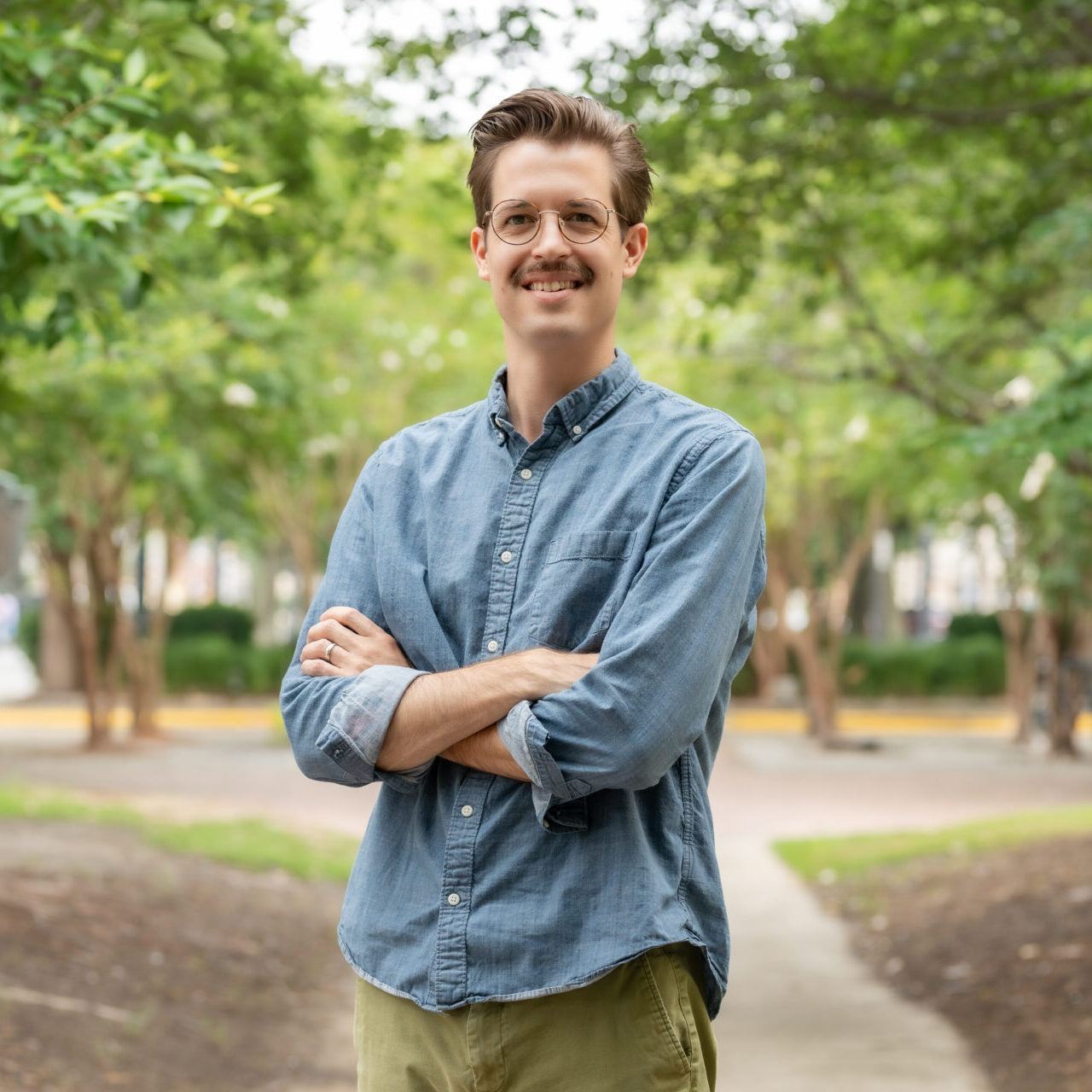 A man with glasses and a mustache stands outside on a path, arms crossed, smiling at the camera. He is wearing a blue button-up shirt and khaki pants, with green trees and a park setting in the background.