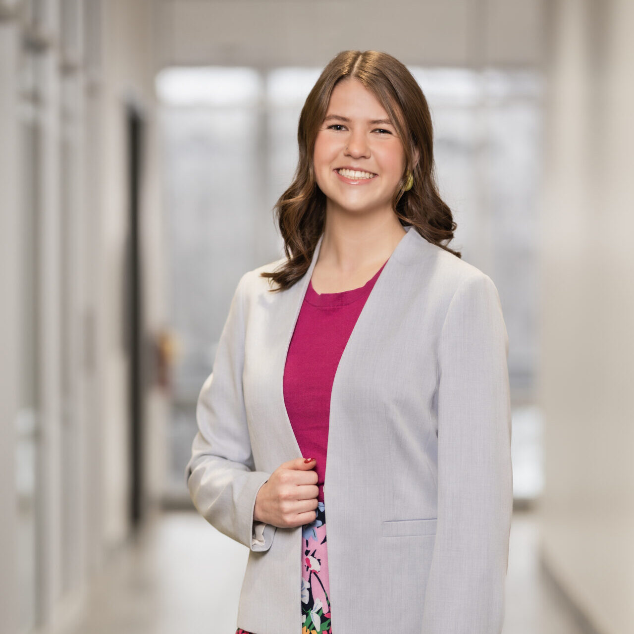 A woman with long brown hair wearing a light gray blazer over a bright pink top stands smiling in a bright modern hallway.