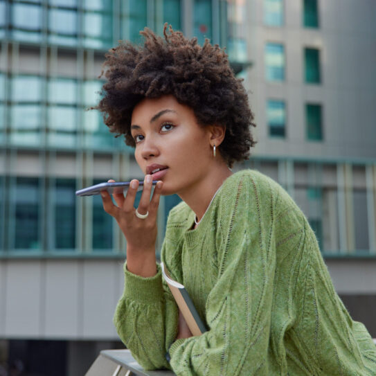 Horizontal shot of thoughtful curly haired woman uses cellphone for communication over speaker records voice message dressed casually poses against modern city building. Modern technologies.