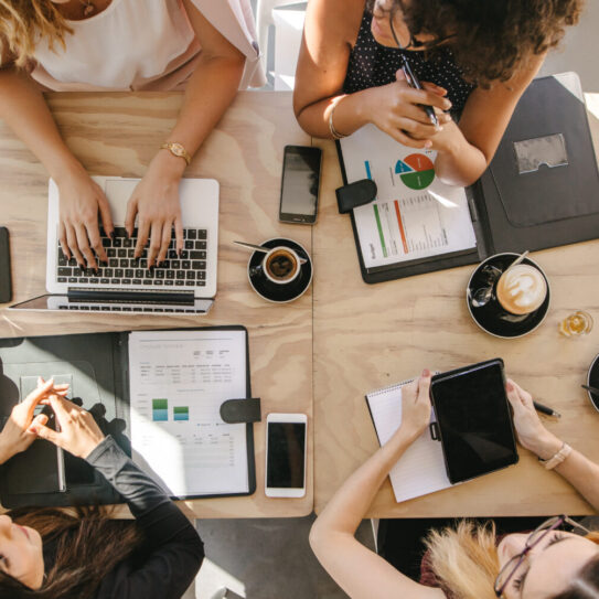 Top view of four women sitting around table in cafe with laptop, digital tablet and documents. Group of women working together in coffee shop.