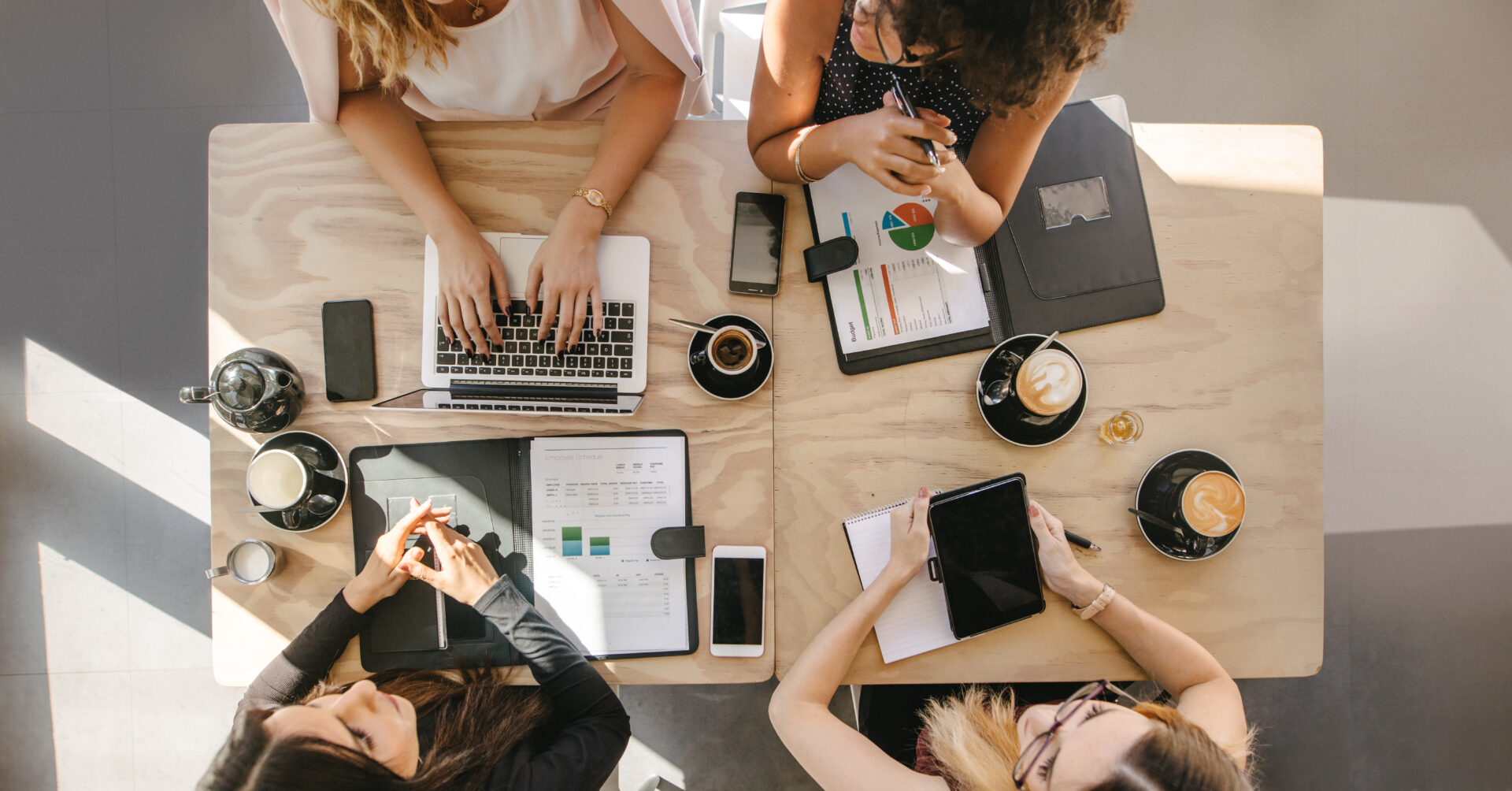 Top view of four women sitting around table in cafe with laptop, digital tablet and documents. Group of women working together in coffee shop.