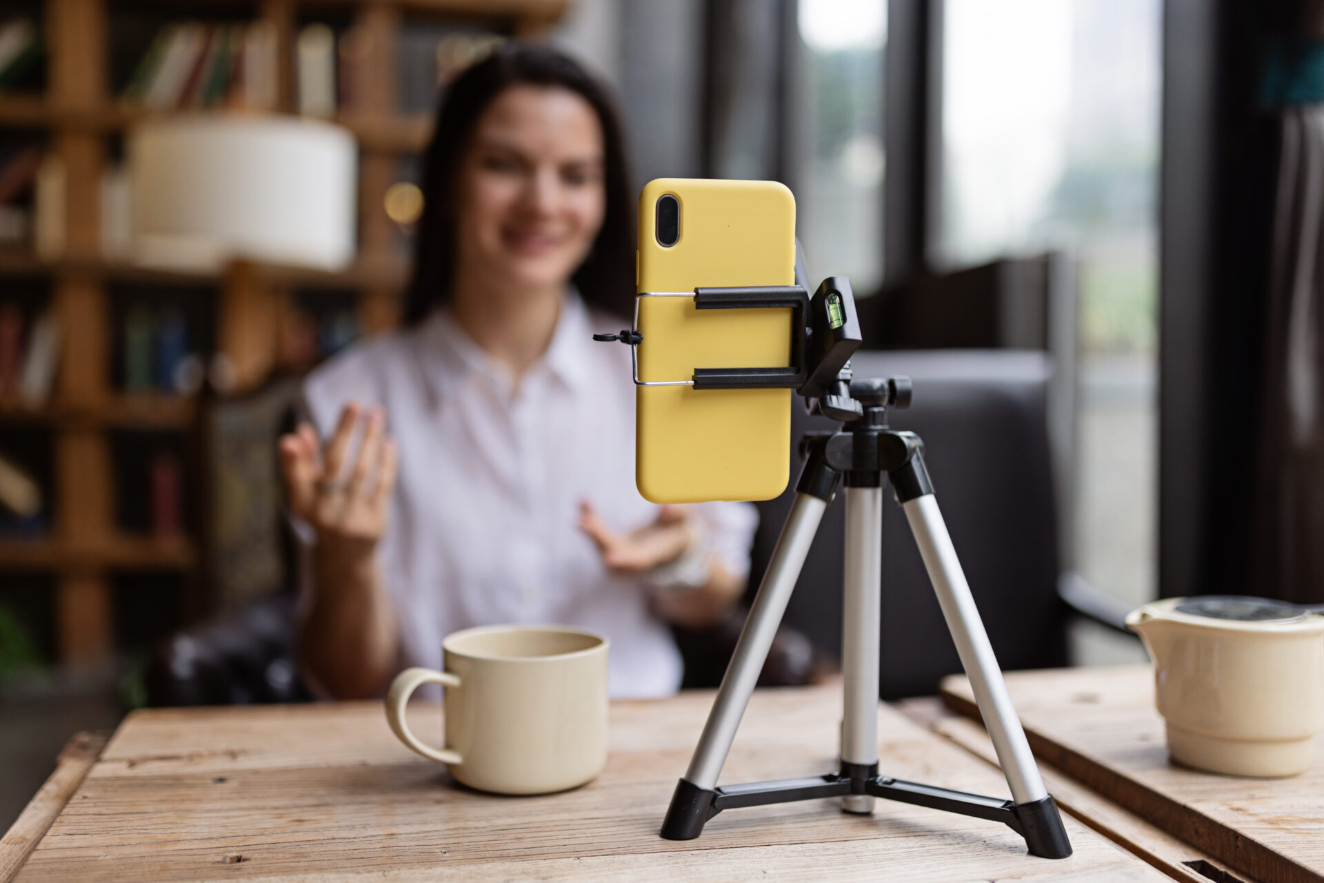 Happy young Caucasian millennial or gen z woman with long brunette hair streaming with smart phone on tripod, shooting social media blog in modern cafe. Influencer using social networks indoor. High quality photo