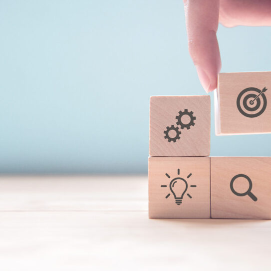 close up of a hand stacking four small wooden cubes. On each cube is an icon representing elements of a marketing plan: gears for the setting phase, a target for the strategy phase, a light bulb for the creation phase, and a magnifying glass for the evaluation phase.