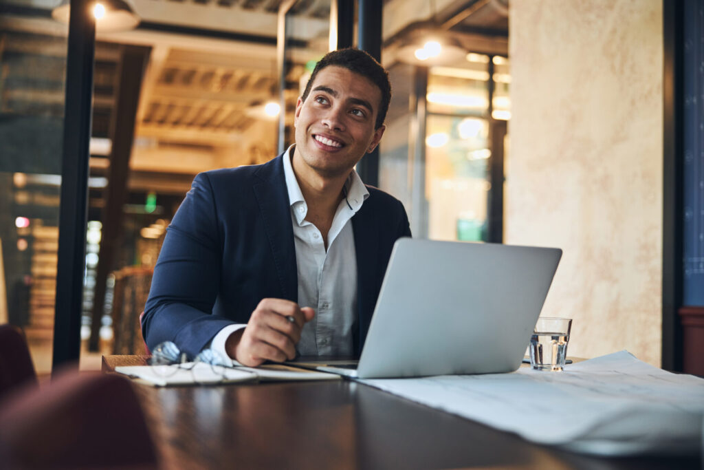 Business man in a conference room sitting in front of a laptop and smiling at someone or something to his right.