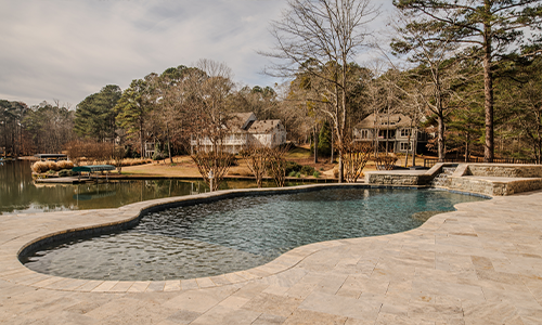 A stone-edged backyard swimming pool overlooks a quiet lake, surrounded by trees and large houses in a serene residential neighborhood.