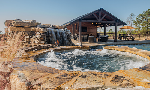 A backyard with a stone hot tub and waterfall next to a swimming pool. In the background, there’s a covered patio with outdoor furniture and an umbrella, surrounded by trees and clear skies.