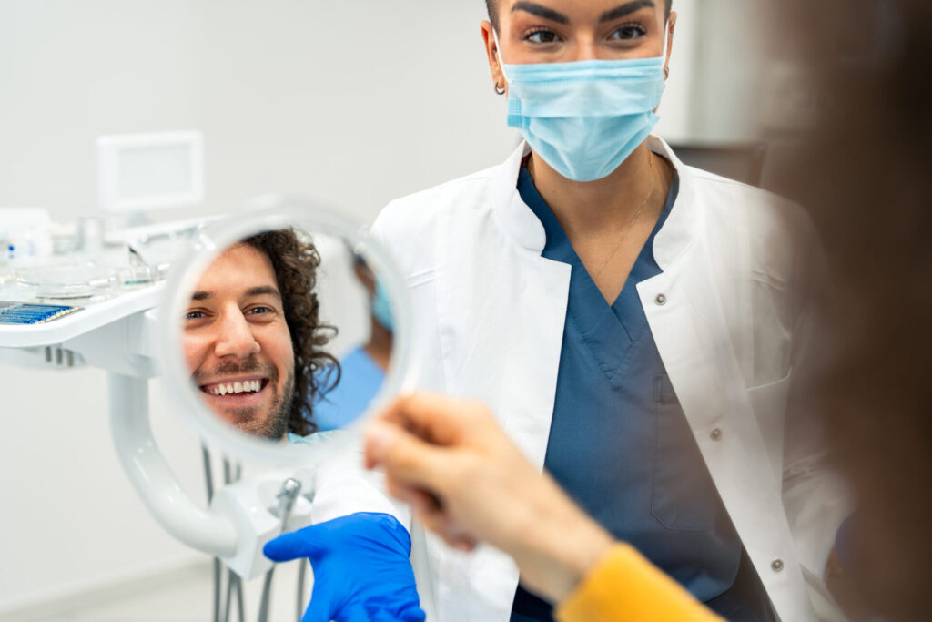 A dentist wearing a mask and gloves stands next to a smiling patient who is looking at their reflection in a handheld mirror during a dental checkup in a bright clinic.