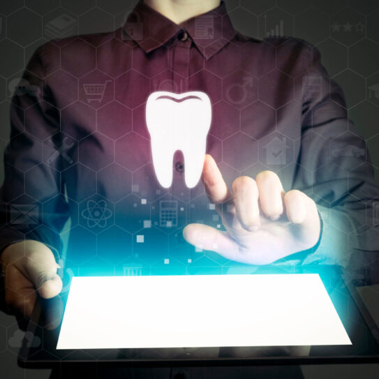 A person in a dark shirt uses a digital tablet, with a glowing tooth icon and virtual interface graphics hovering above the screen, symbolizing digital dental technology.