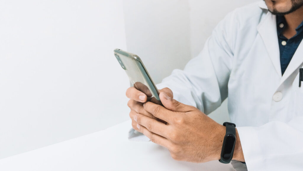A dentist wearing a white lab coat and a black wristband is sitting at a desk, holding and using a smartphone. The person's face is partially out of frame.