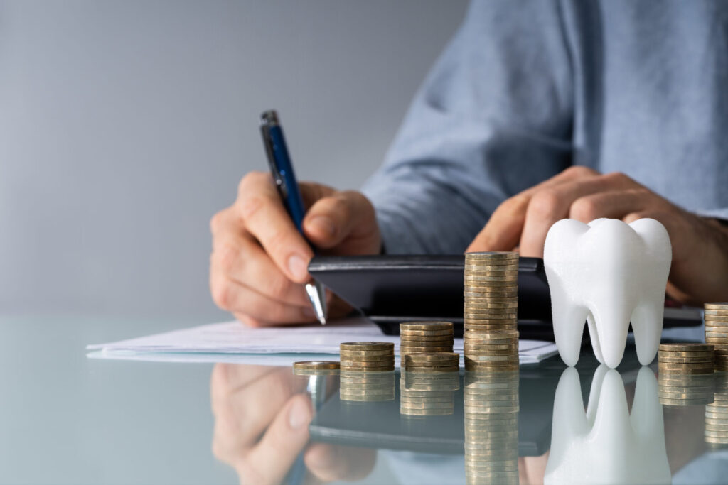 A person calculates finances with a pen and calculator beside stacked coins and a model tooth, symbolizing dental expenses or dental insurance costs.