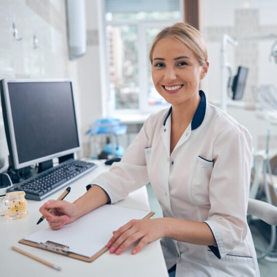 A smiling woman in a white lab coat sits at a desk with a clipboard, pen, and computer in a bright dental office. Dental equipment and a patient chair are visible in the background.