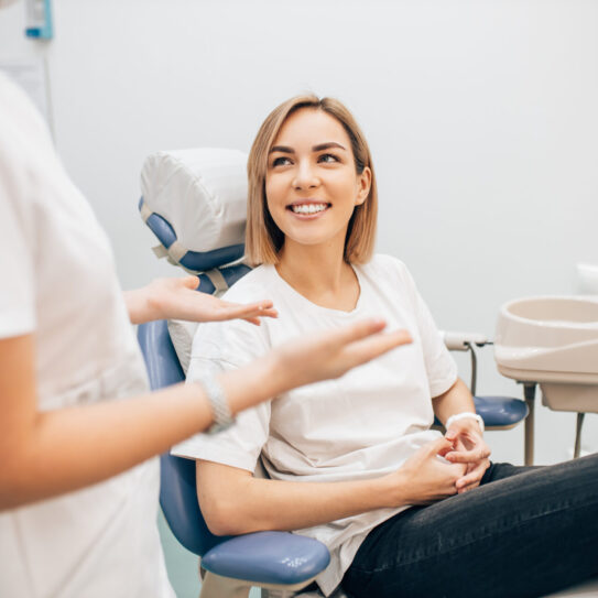 A woman sits in a dental chair smiling at a dental professional who is standing and gesturing with her hands. The setting is a clean, modern dental office.