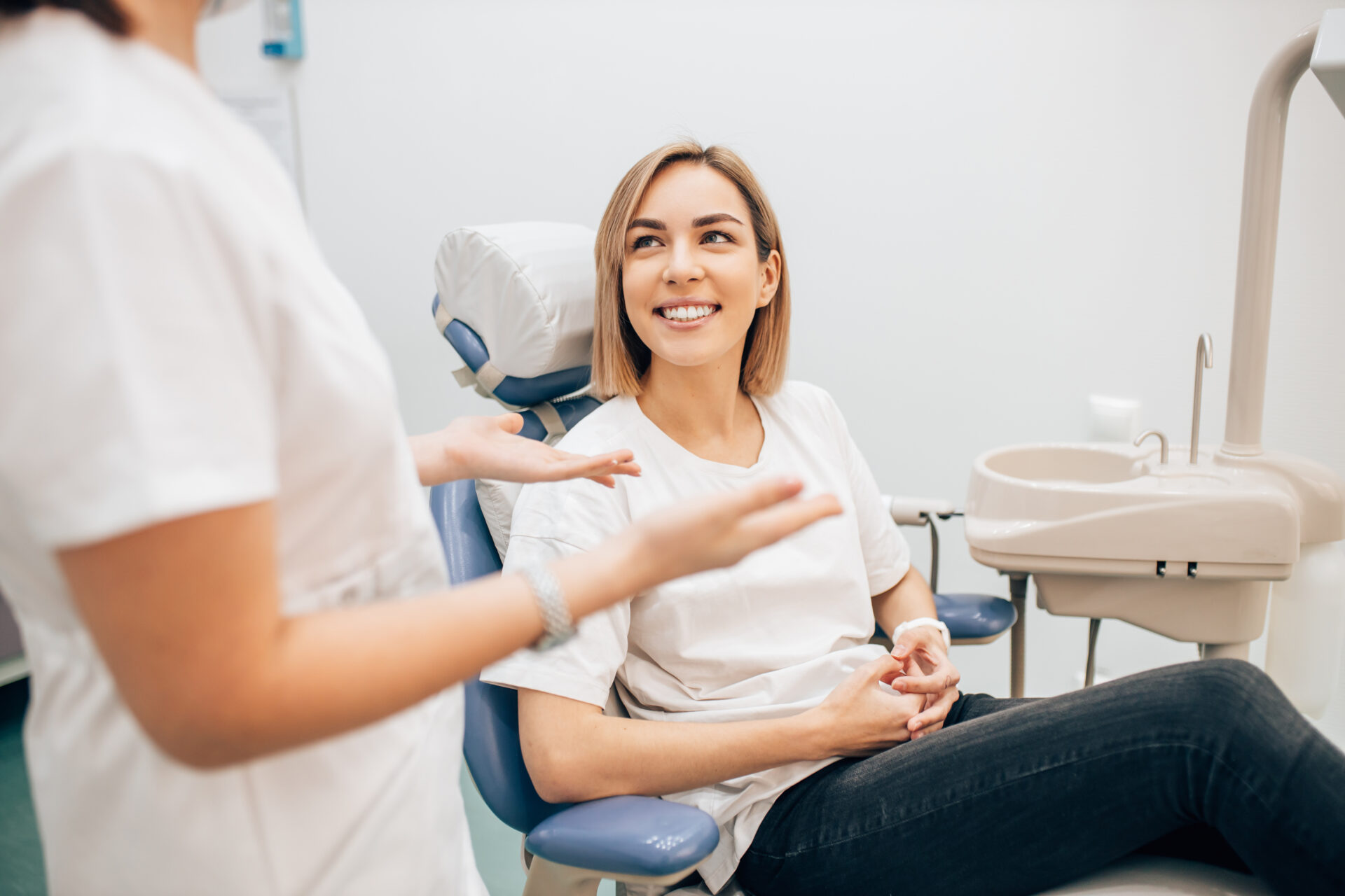 A woman sits in a dental chair smiling at a dental professional who is standing and gesturing with her hands. The setting is a clean, modern dental office.