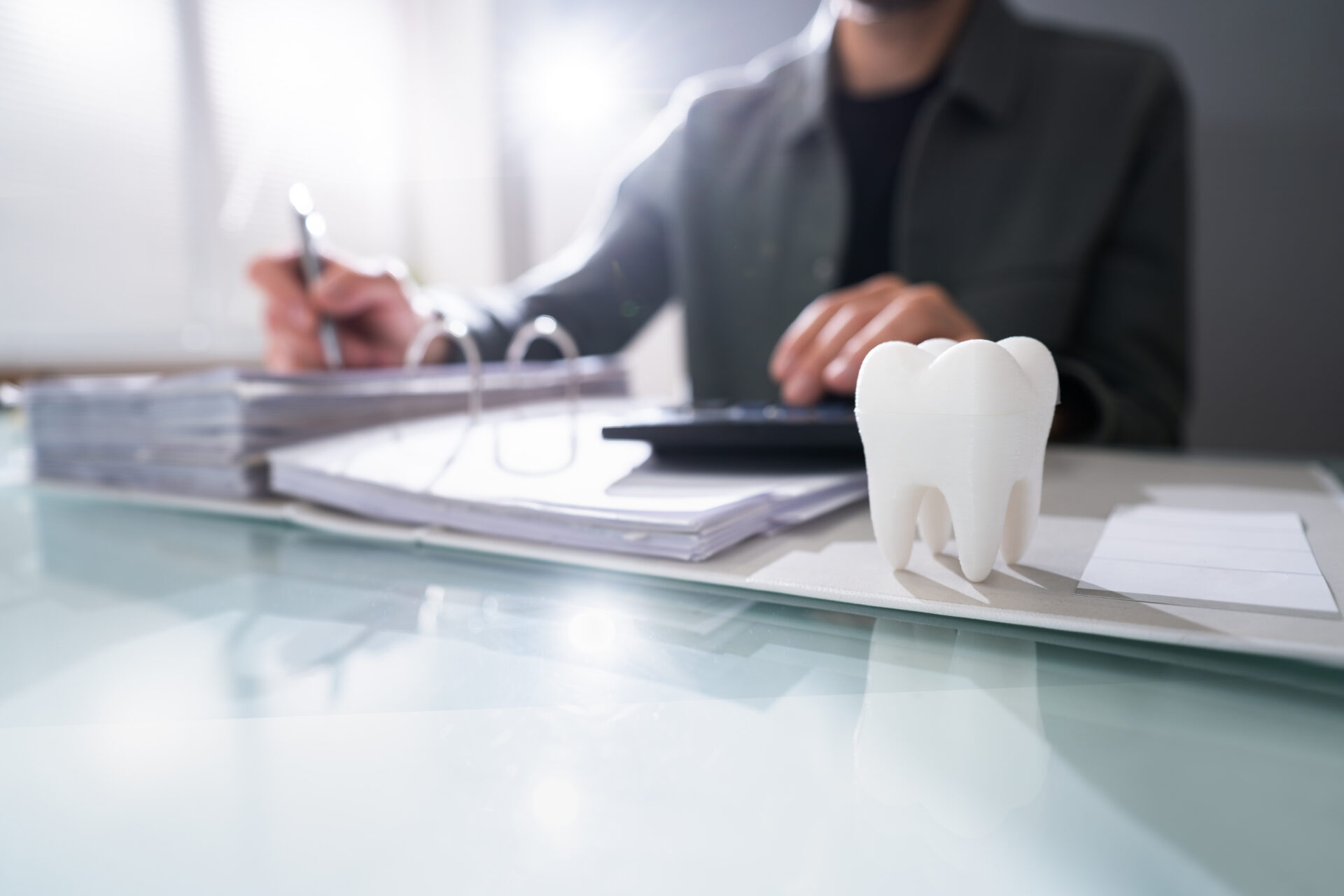 A person writes at a desk with paperwork and a calculator. In the foreground, there is a large model of a tooth placed on the glass surface.