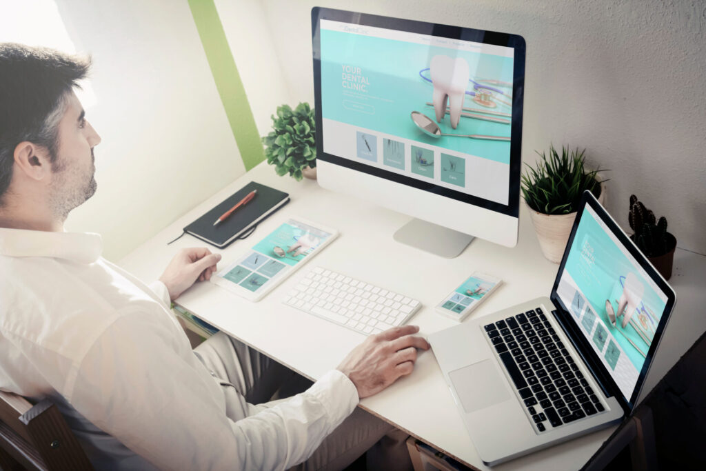 A person sits at a white desk using a desktop computer, laptop, tablet, and smartphone, all displaying the same dental clinic website. The desk has a keyboard, notepad, pen, potted plants, and mouse.