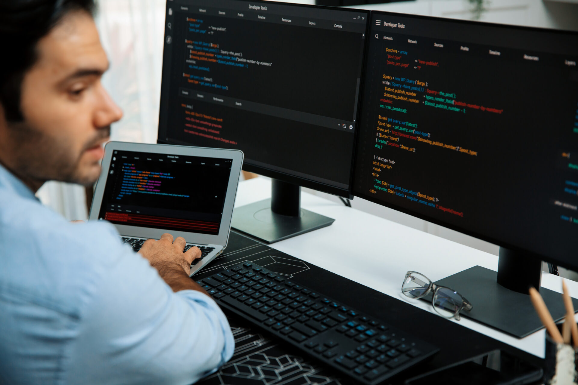 A man sitting at a desk is coding on a laptop, with two external monitors displaying lines of colorful code. A keyboard, mouse, and glasses are on the desk next to him.