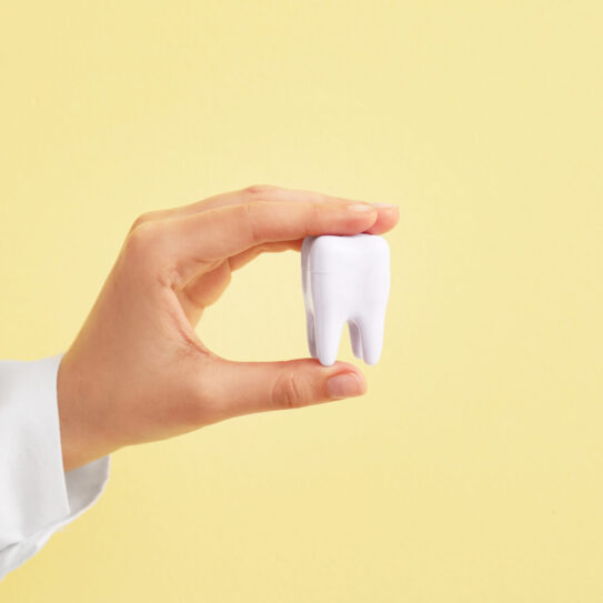 A hand in a white sleeve holds a large white model tooth against a pale yellow background.