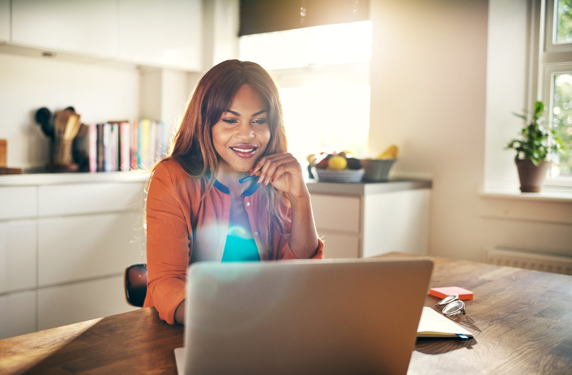A woman sits at a kitchen table, smiling as she looks at a laptop screen. Sunlight streams through the window, and kitchen items and books are visible in the background.