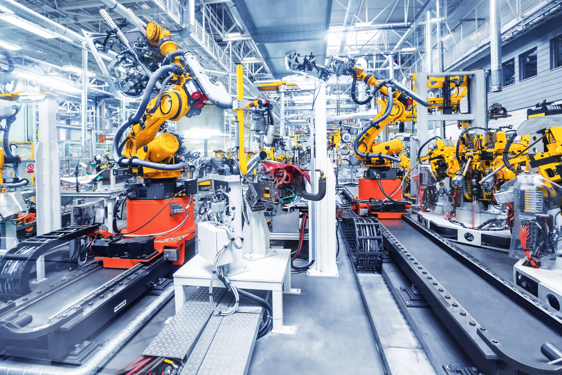 Bright, modern automotive factory filled with yellow robotic arms assembling car parts on production lines, surrounded by metal structures and overhead lights.